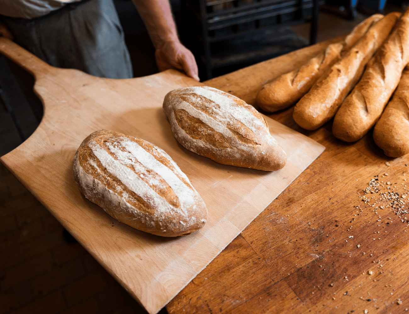 Bäcker mit einer Holzschaufel, auf der zwei frisch gebackene, mit Mehl bestäubte Rundbrote liegen; mehrere Baguettes liegen auf einem Holztisch.