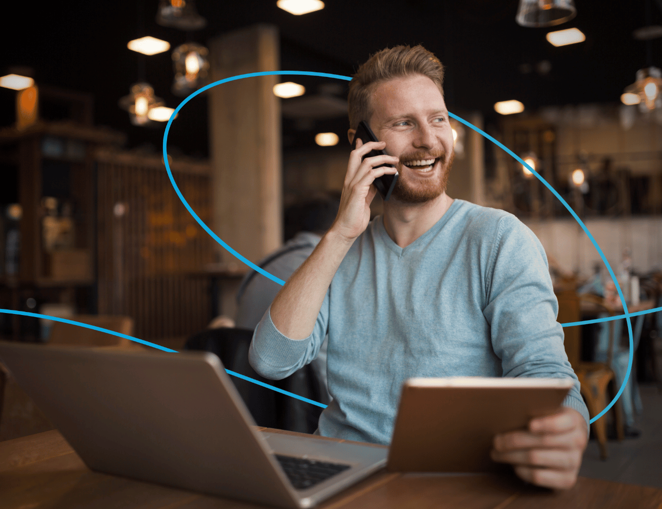 Ein lächelnder Mann in einem blauen Pullover telefoniert, hält ein Tablet in der Hand und sitzt an einem Tisch mit einem Laptop in einem gemütlichen Café.