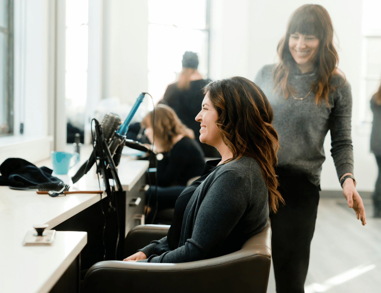 Eine Frau sitzt lächelnd in einem Friseurstuhl, während eine Stylistin neben ihr steht. Beide wirken glücklich in einem hellen, modernen Salon.