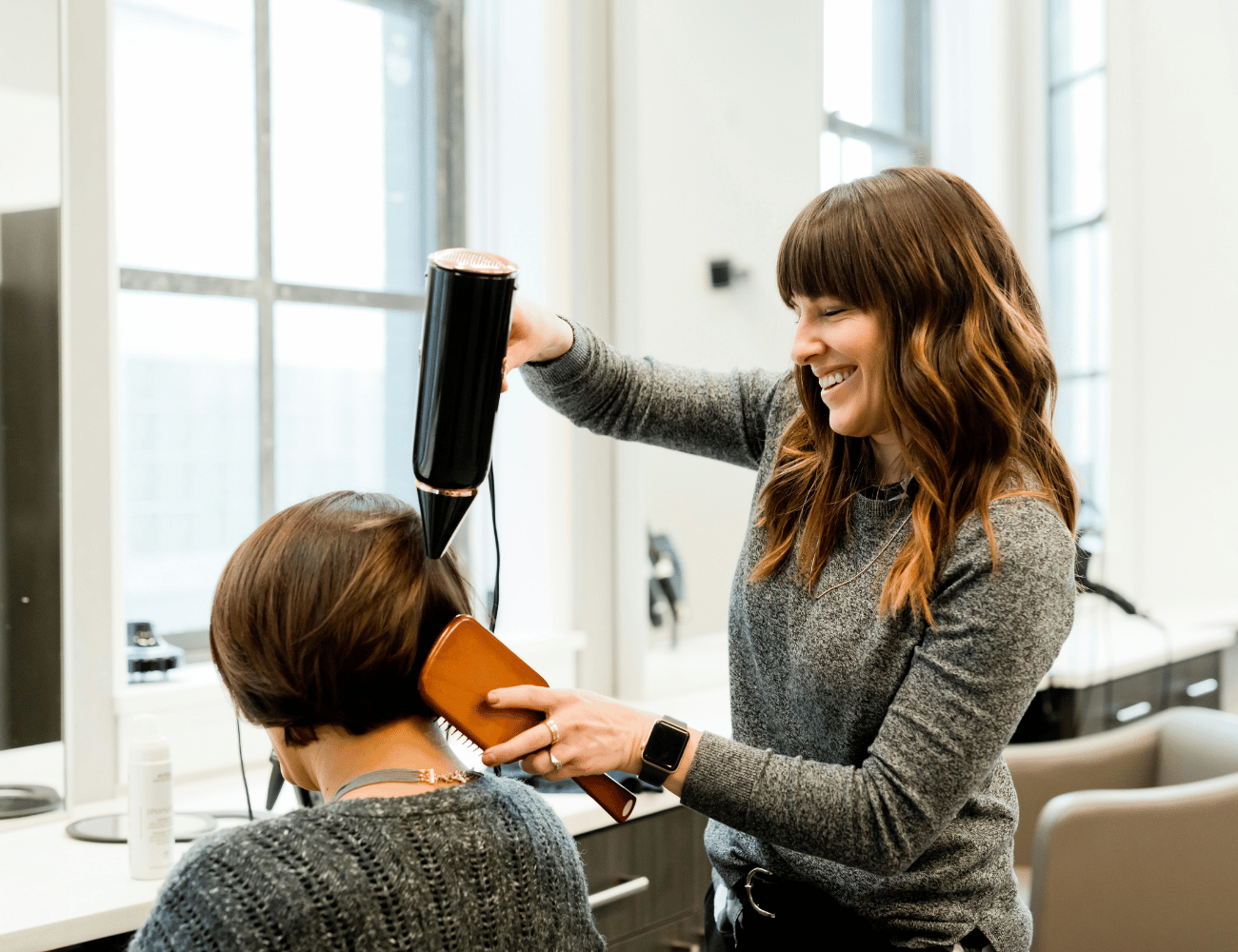 Eine Friseurin föhnt das kurze Haar einer Kundin in einem hellen Salon, beide lächeln und sind konzentriert bei der Arbeit, Styling-Werkzeuge und -Produkte sind auf dem Tresen zu sehen.