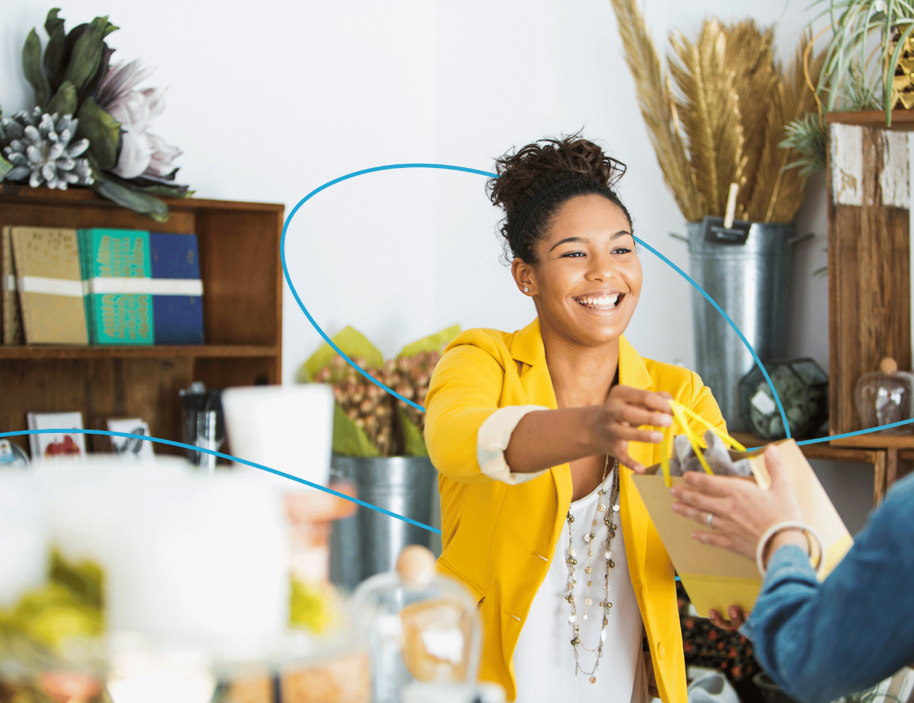 Eine lächelnde Frau in einer gelben Jacke reicht einem Kunden in einem gemütlichen, dekorierten Laden eine Einkaufstüte.