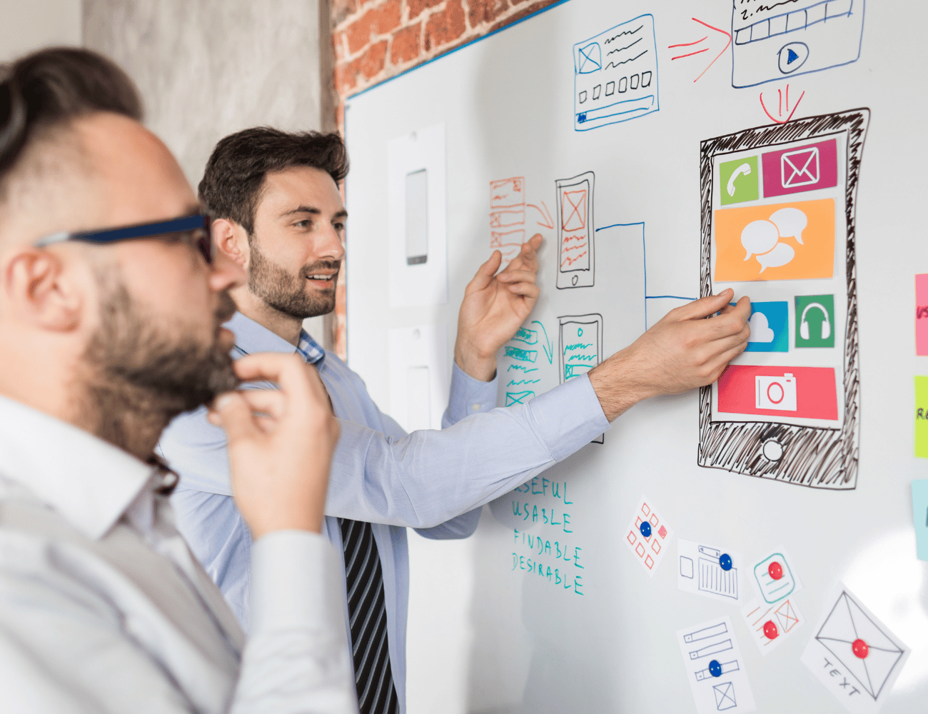Zwei Männer arbeiten gemeinsam an Entwürfen für mobile Apps und analysieren Skizzen und Wireframes auf einem Whiteboard. Einer von ihnen deutet auf ein großes Smartphone-Layout.