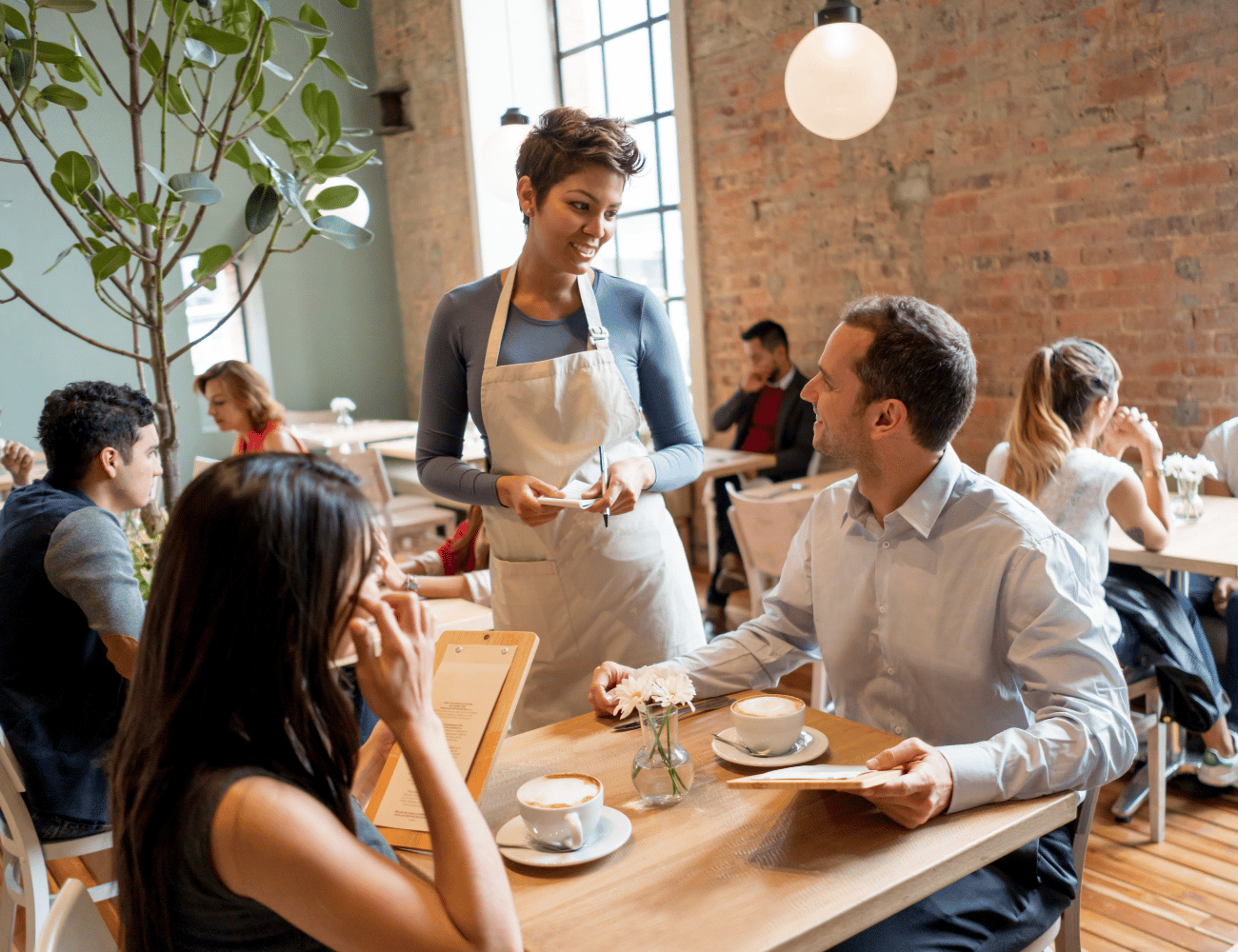 Eine Kellnerin nimmt die Bestellung eines Paares entgegen, das in einem gemütlichen Café mit Backsteinwänden sitzt. Im Hintergrund sind weitere Gäste beim Essen zu sehen.