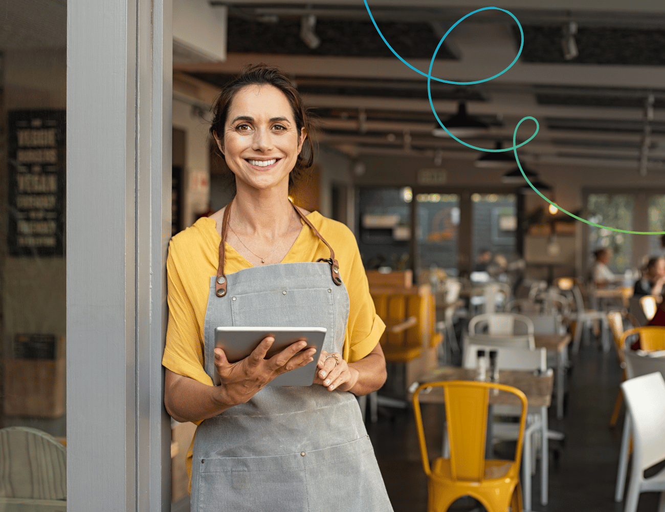 Lächelnde Frau in einem gelben Hemd und einer Schürze hält ein Tablet in der Hand und steht am Eingang eines Cafés mit gelben und weißen Stühlen im Inneren.