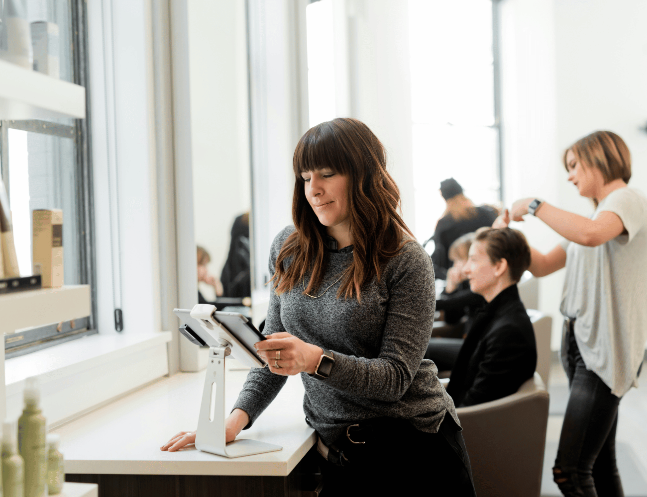 Frau benutzt ein Tablet an einem Tisch in einem Friseursalon, während im Hintergrund eine andere Friseurin die kurzen Haare einer Person stylt. Helles, modernes Interieur.