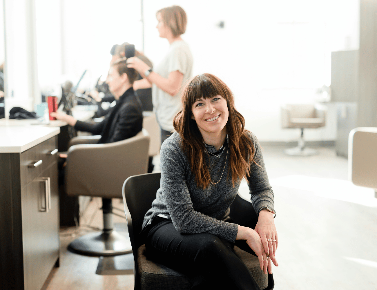 Frau lächelt und sitzt in einem Friseurstuhl, im Hintergrund arbeiten Stylist:innen an Kund:innen. Helles, luftiges Interieur.