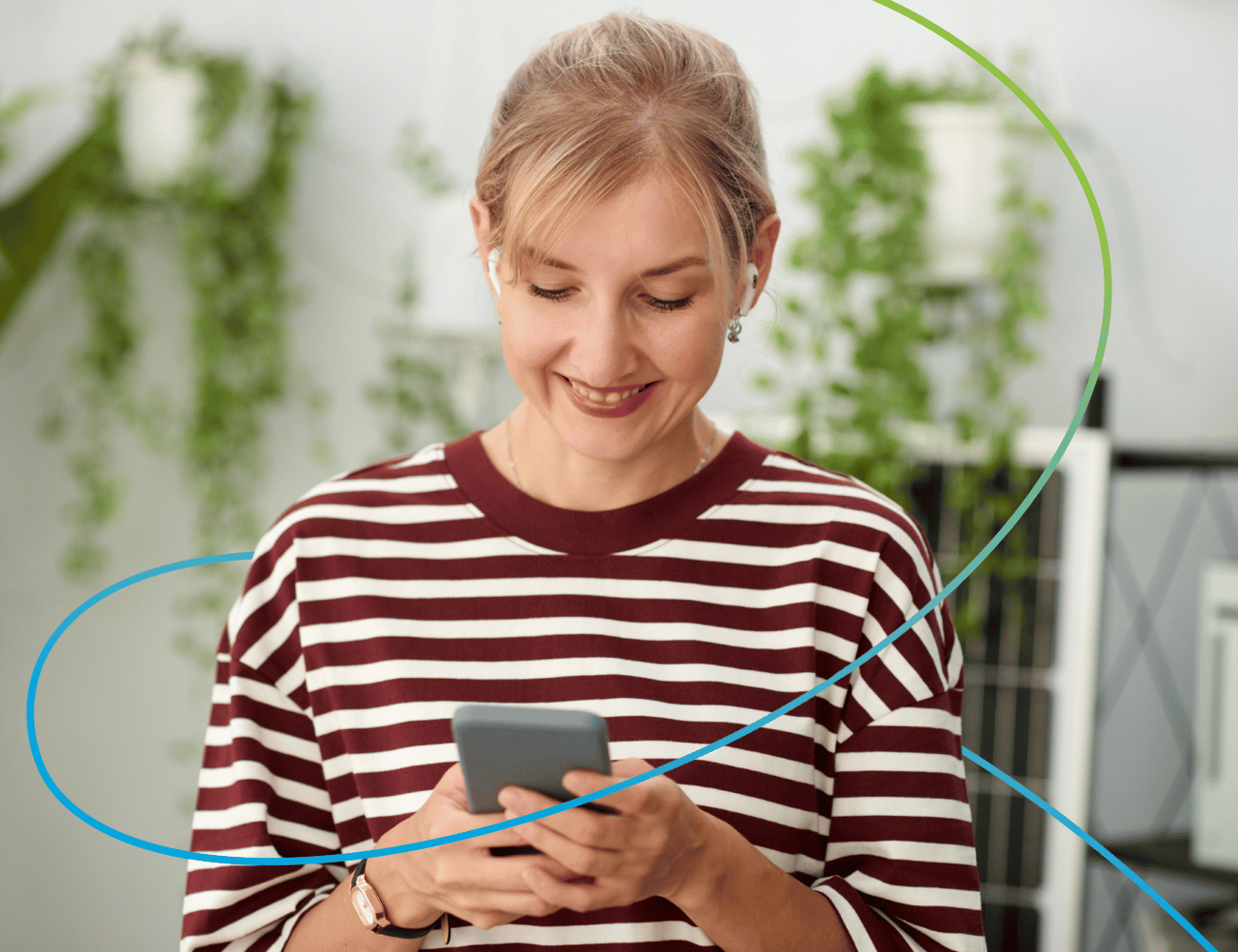 Lächelnde Frau in einem kastanienbraun-weiß gestreiften Shirt, die auf ihr Smartphone schaut, verschwommene Topfpflanzen im Hintergrund.