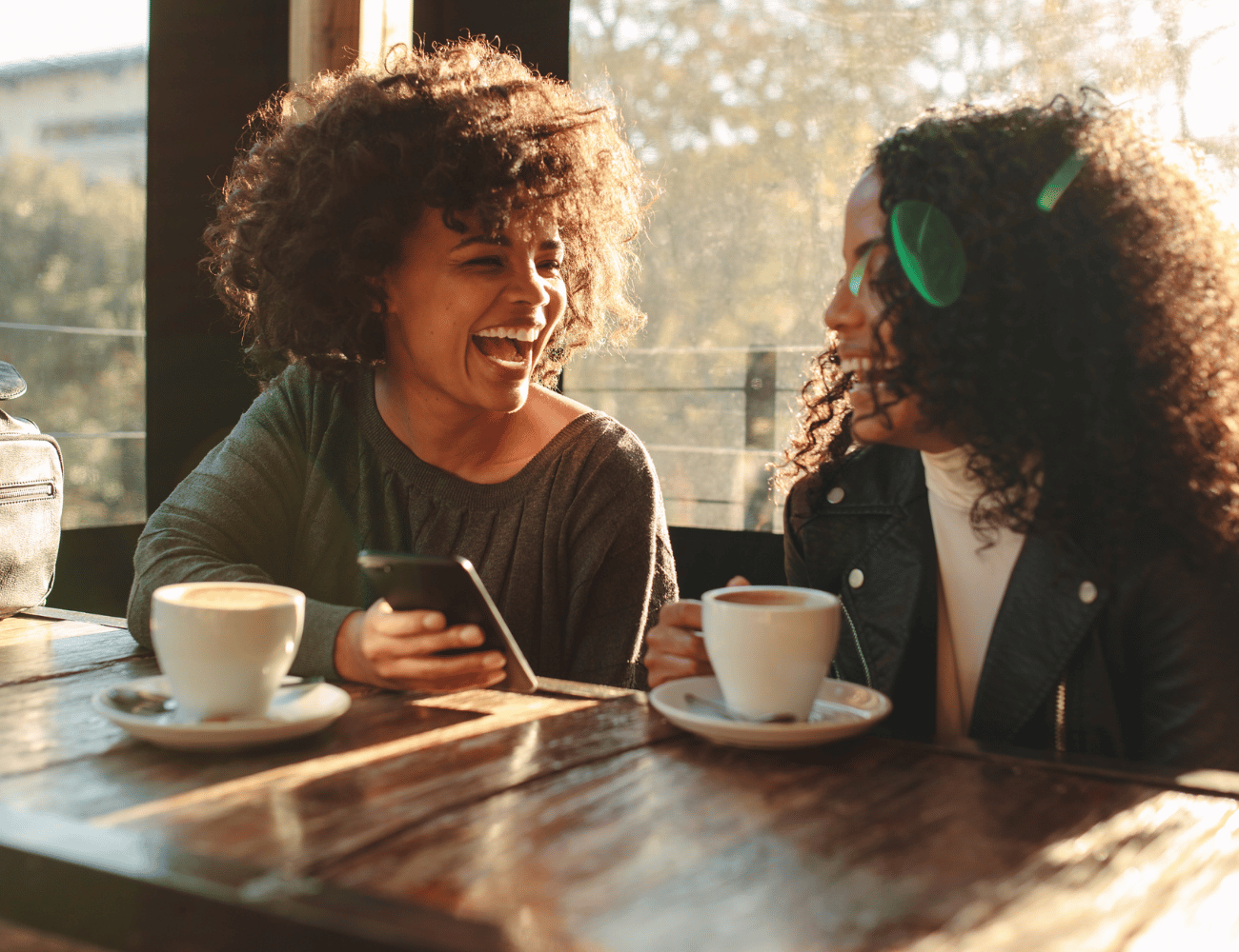 Zwei Personen sitzen an einem Holztisch in einem Café, lachen und halten Kaffeetassen in den Händen, während Sonnenlicht durch das Fenster scheint.