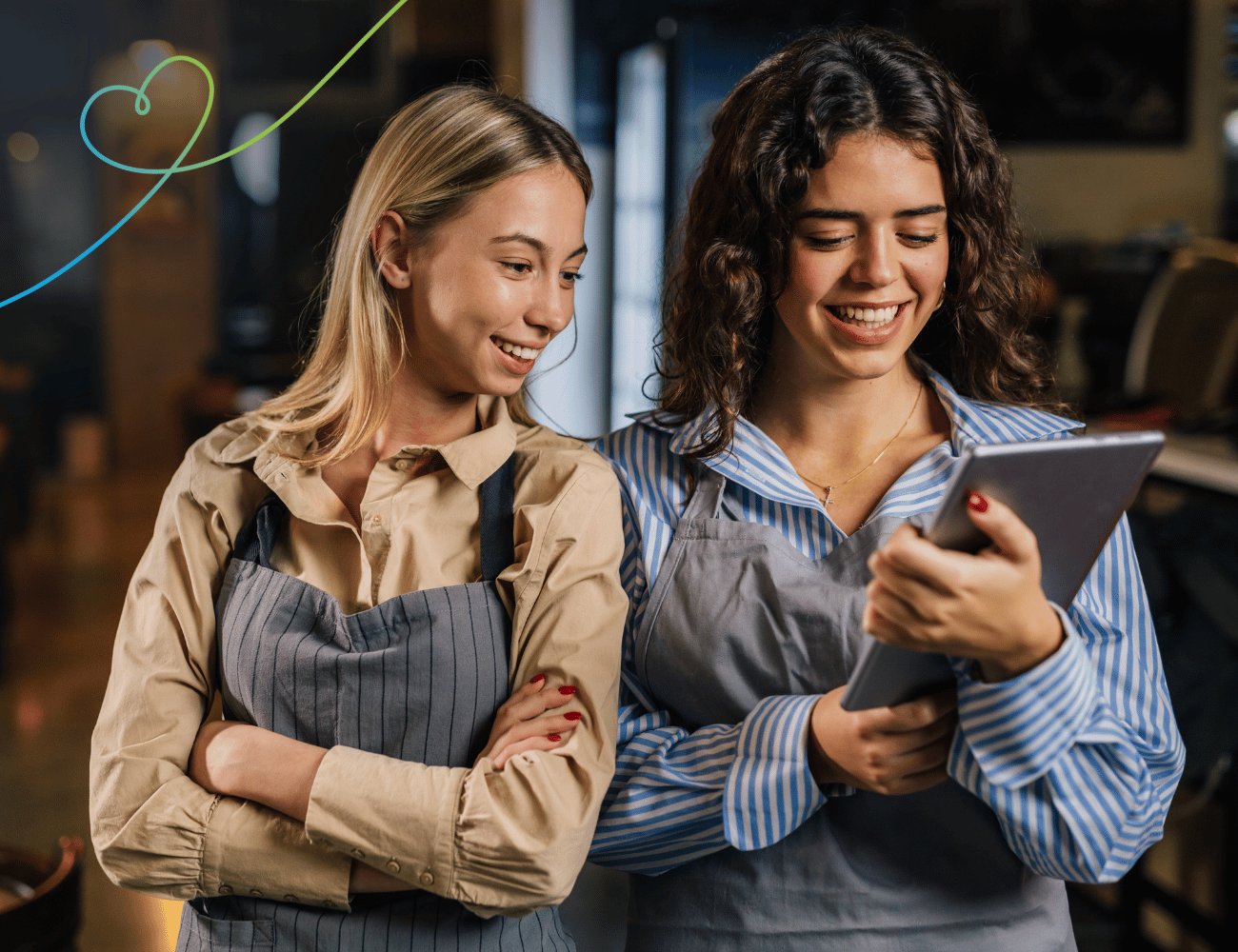 Zwei Frauen in Schürzen stehen zusammen und schauen lächelnd auf ein Tablet. Sie scheinen sich in einer gemütlichen, warm beleuchteten Umgebung zu befinden.