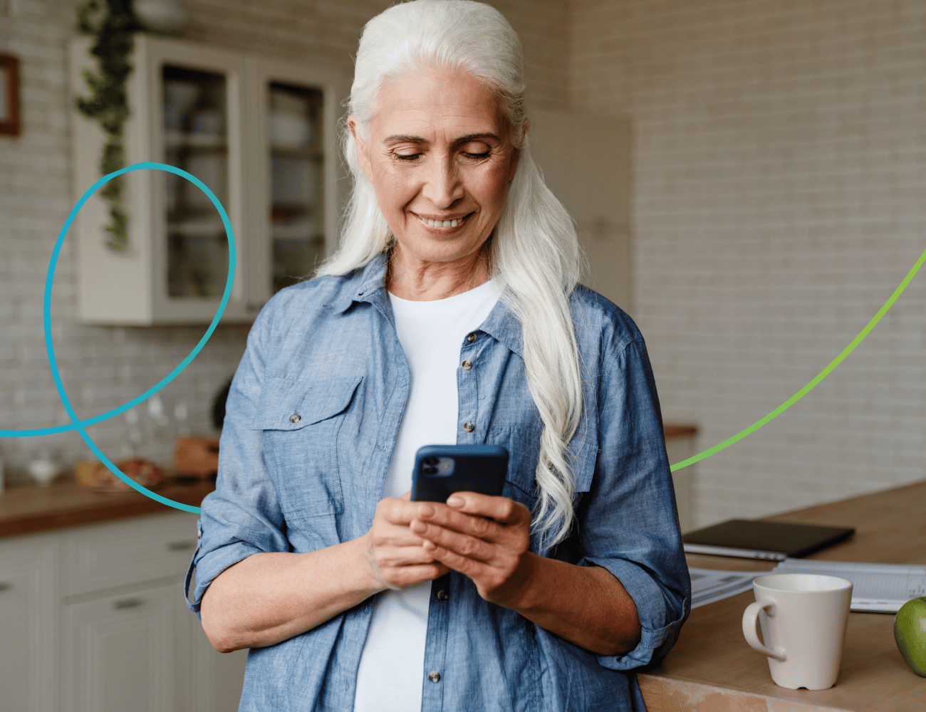 Ältere Frau mit langen weißen Haaren lächelt, während sie in einer Küche ein Smartphone benutzt. Sie trägt ein Jeanshemd. Auf der Arbeitsplatte stehen eine Tasse und ein Laptop.