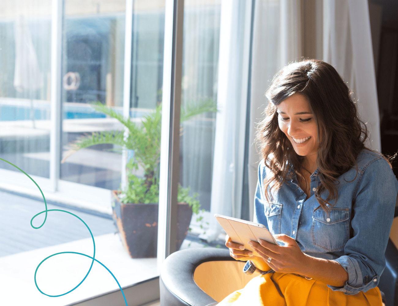 Frau in Jeanshemd lächelt, während sie ein Tablet in der Nähe eines Fensters mit einer Topfpflanze und transparenten Vorhängen im Hintergrund benutzt.