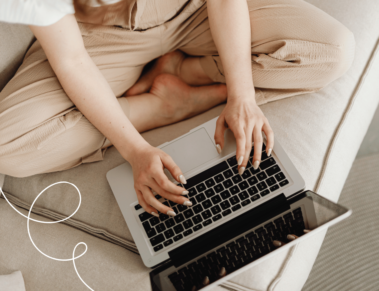 Person, die mit gekreuzten Beinen auf einer Couch sitzt und auf einem Laptop tippt, der auf ihrem Schoß liegt, mit neutraler Kleidung.
