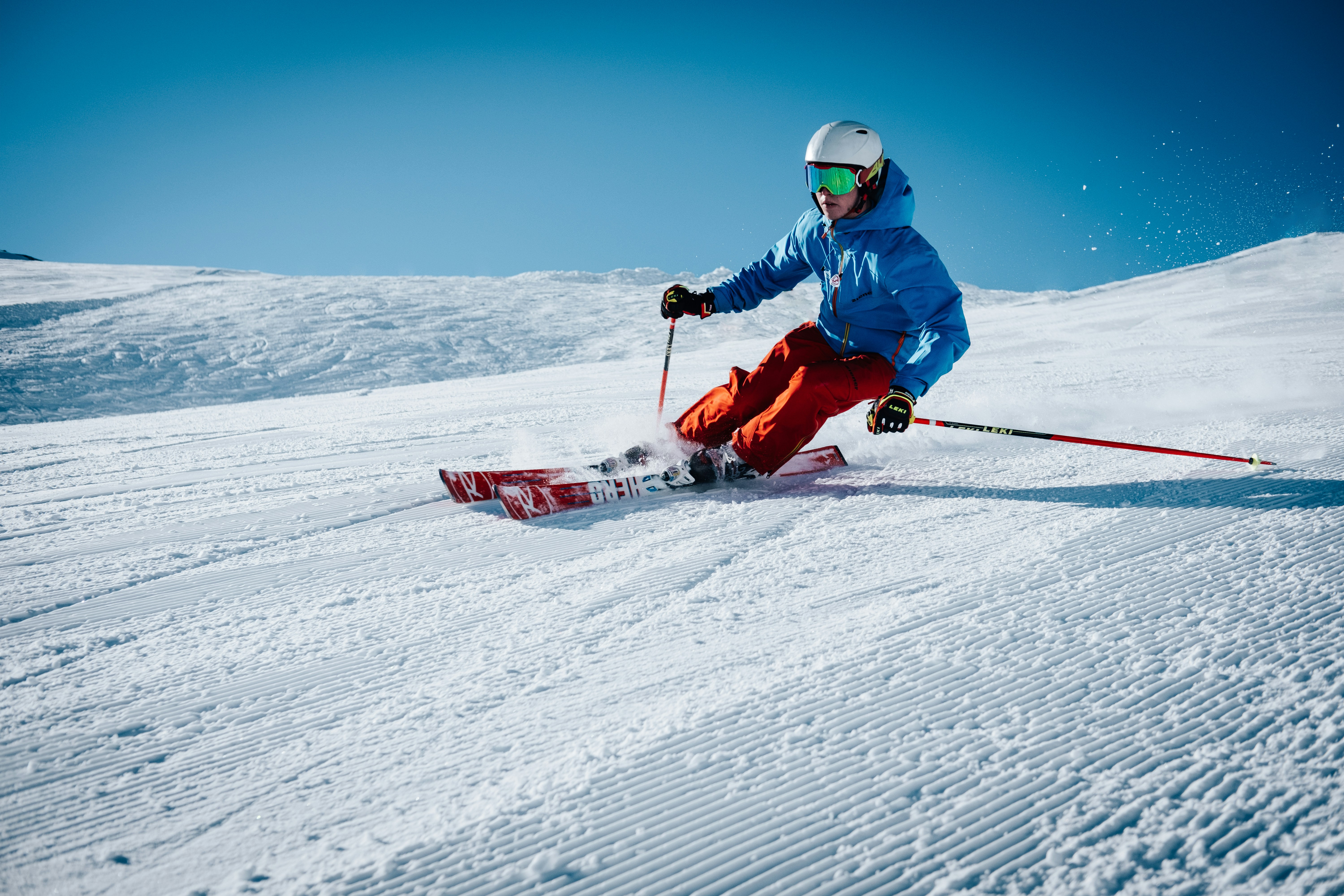 Ein Skifahrer in blauer Jacke und roter Hose gleitet eine verschneite Piste hinunter unter klarem blauem Himmel und hinterlässt Spuren im Schnee.