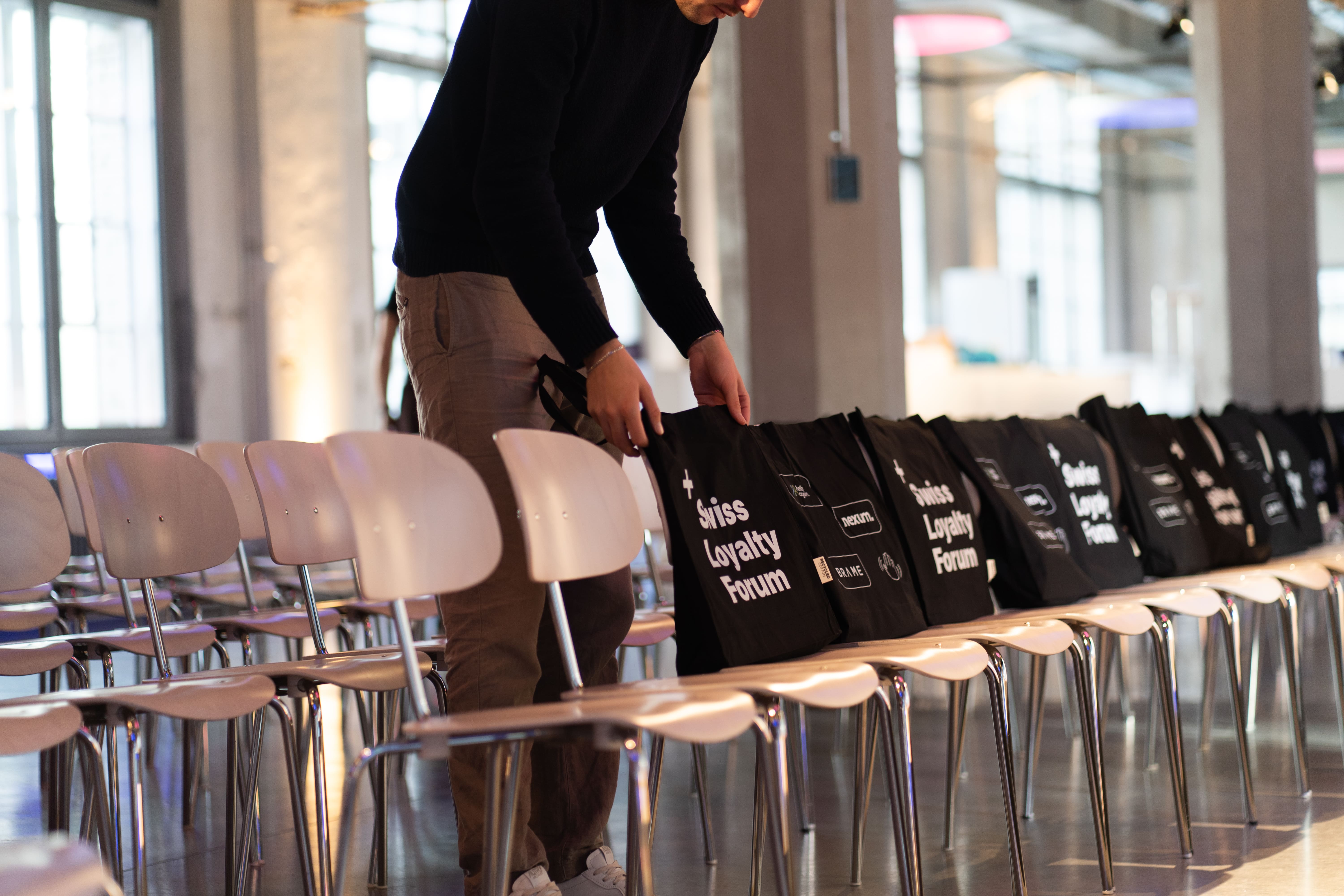 Person, die schwarze Tragetaschen auf Stühlen in einem Konferenzraum mit großen Fenstern und Einrichtung im Industriestil ordnet.