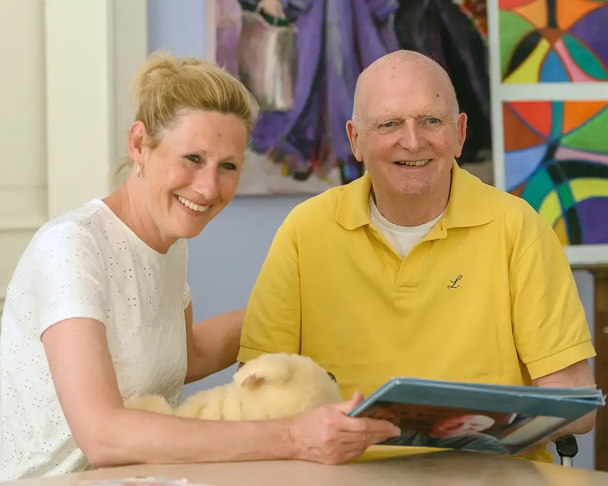 A smiling woman and man sit at a table, holding a book with a small dog. Colorful artwork is displayed in the background.