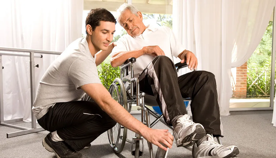 un auxiliaire de vie aide une personne en situation de handicap moteur à s'installer dans son fauteuil roulant