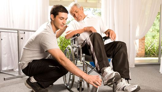 un auxiliaire de vie aide une personne en situation de handicap moteur à s'installer dans son fauteuil roulant