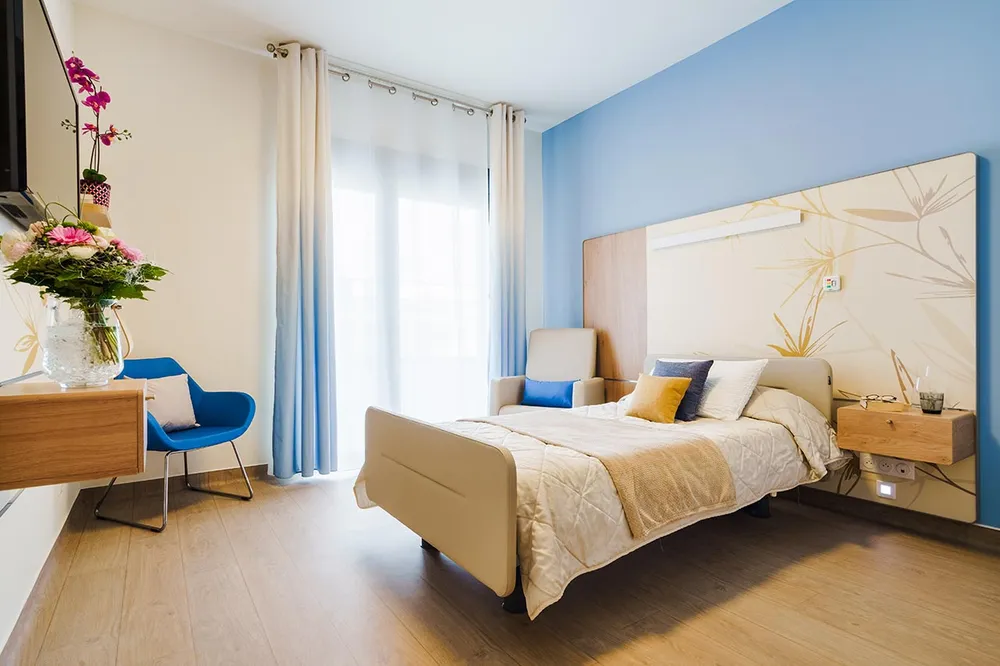 Chambre maison retraite bleu blanc cocooning moderne lumineuse grande fenetre avec rideau assiortie fauteil bleu mer avec table et orchidées