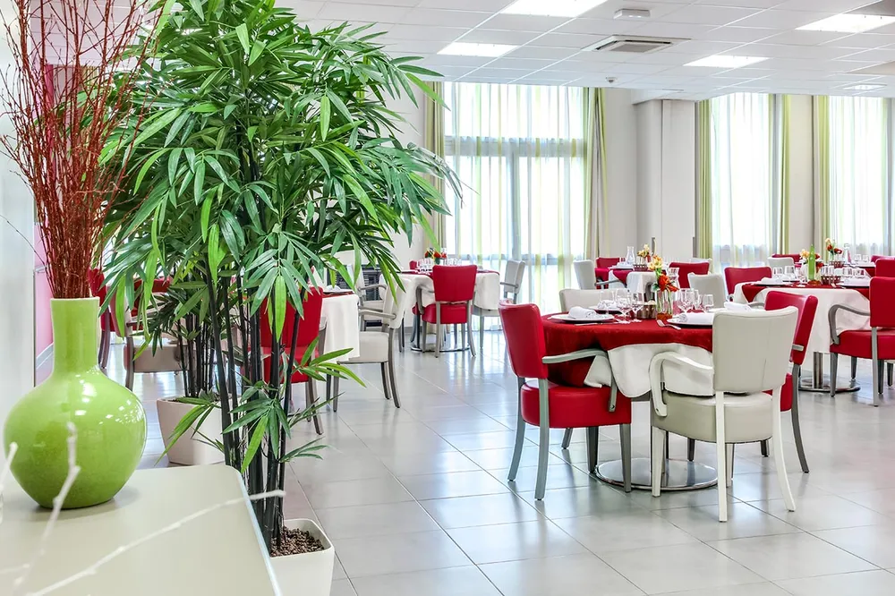 Salle lumineuse avec bambou et baie vitrée, tables rondes rouges et dressées avec fauteils rouges et beiges