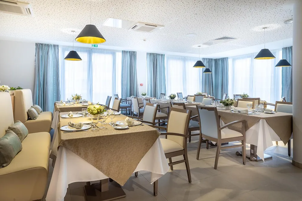 Salle de restaurant lumineuse Pavillon des Arts à Saint-Brice-sous-Foret baie vitrée rideaux bleu gris, tables carrées dorées dressées
