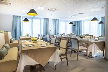 Salle de restaurant lumineuse Pavillon des Arts à Saint-Brice-sous-Foret baie vitrée rideaux bleu gris, tables carrées dorées dressées