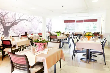 Salle de restaurant avec tables carrées dressées avec bouquet fleurs frais, mur sakura japonais et lumière naturel fenêtre exposition sud