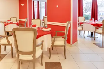 Salle de restaurant avec tables rondes décorées et chaises beiges et rouges, murs rouges, vue sur jardin et nature