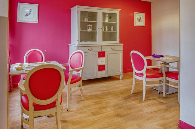 grande salle de jeux avec tables carrées et fauteils rouges et placard avec statuettes blanc mur rouge