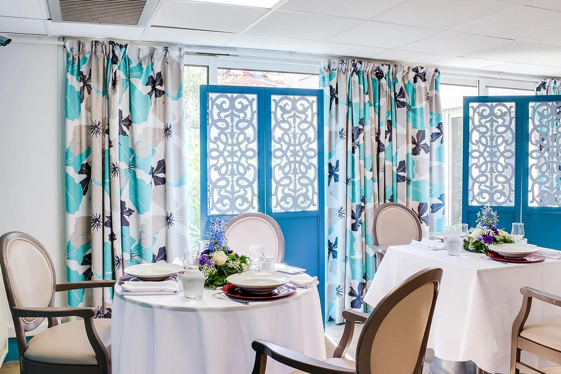 Salle de restaurant lumineux avec tables dressées, rideaux florales, ehpad bleu soleil à Nice