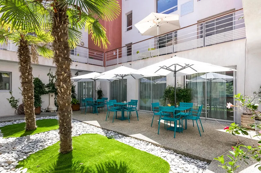 Patio extérieur avec tables et chaises turquoise, palmiers, parasols blanc, EHPAD Palmeraie à Nice
