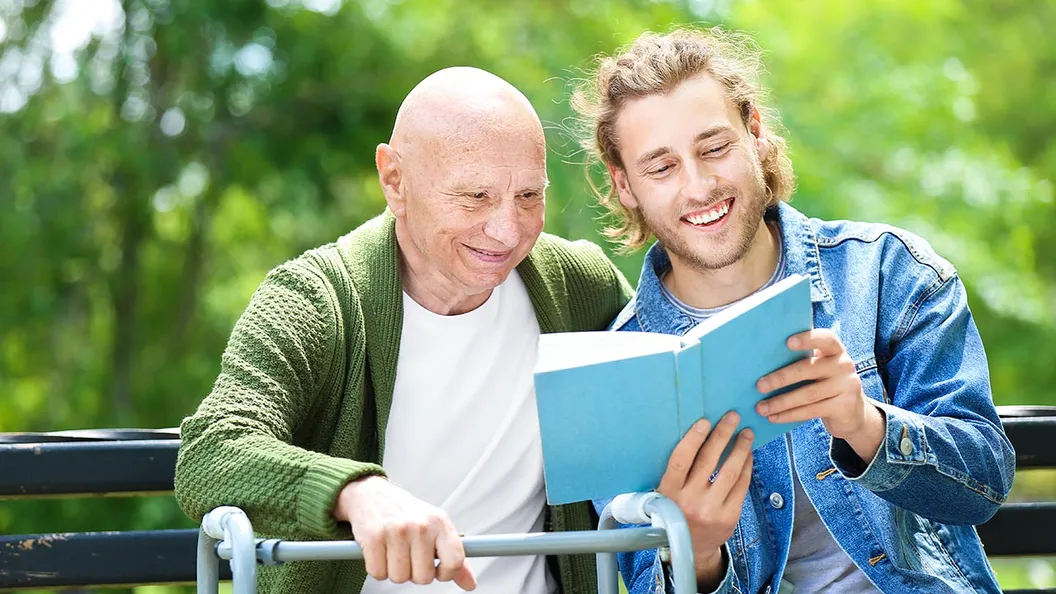 une homme âgé et un homme plus jeune sont assis sur un banc à l'extérieur, le plus jeune montre un livre au plus âgé