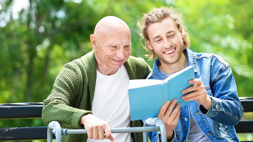 une homme âgé et un homme plus jeune sont assis sur un banc à l'extérieur, le plus jeune montre un livre au plus âgé