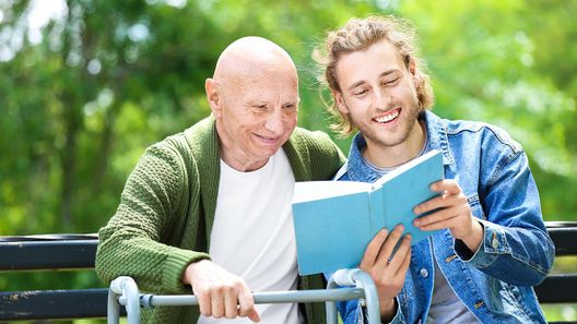 une homme âgé et un homme plus jeune sont assis sur un banc à l'extérieur, le plus jeune montre un livre au plus âgé
