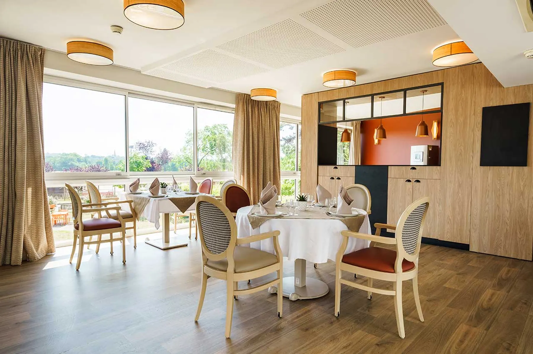 Grande salle dinatoire avec meubles en bois, vue panoramique sur la ville et la nature, tables restaurant dressées et décorées avec une belle exposition soleil et décoration chic moderne