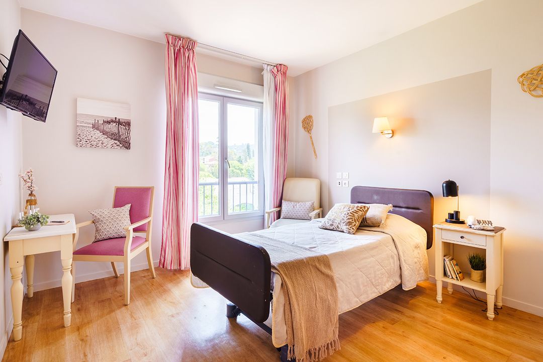 Chambre lumineuse avec lit simple, rideaux roses, télévision murale, bureau avec chaise maison retraite le cannet