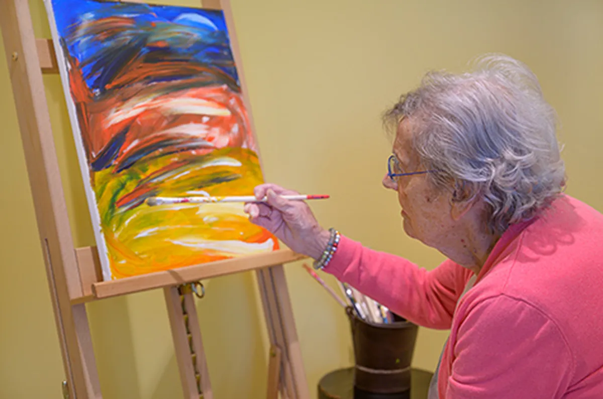 Oudere persoon schildert abstract landschap op canvas in goed verlichte kamer. Creativiteit is cruciaal in de dementiezorg.
