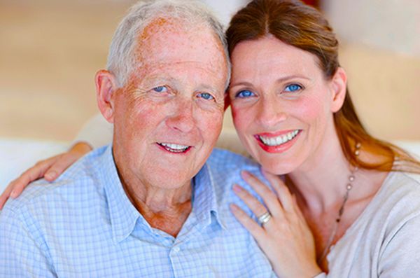 Vader en dochter glimlachen naar de camera, arm om schouder. Warme en steunende familierelaties zijn essentieel in de dementiezorg.