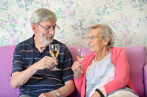 Ouder echtpaar lacht en proost met wijnglazen op een paarse bank. Een knusse setting die belangrijk is voor de levenskwaliteit in de dementiezorg.