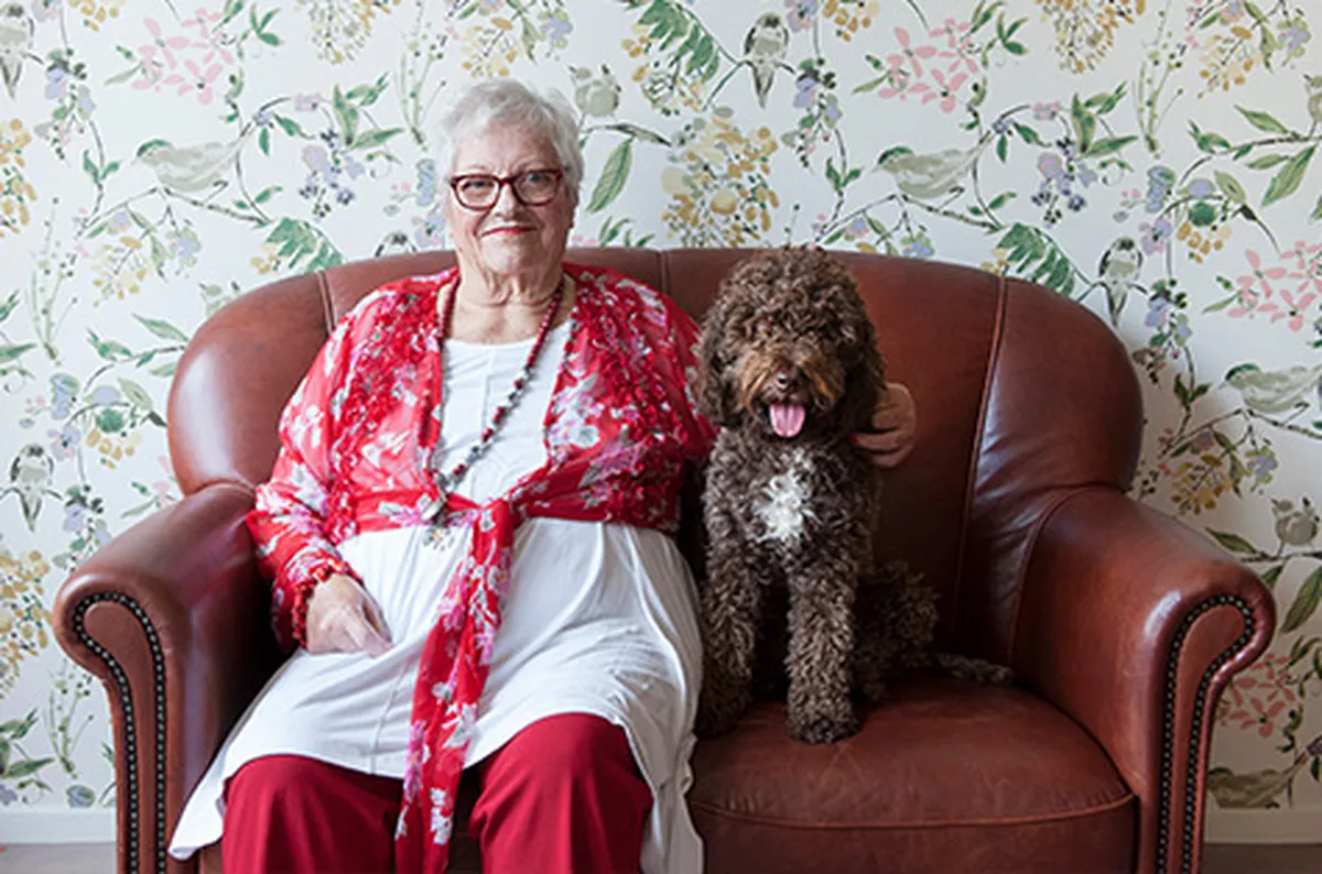 Oudere vrouw in rood-witte kleding op een leren bank met een gekrulde hond, tegen bloemenbehang. De troost van huisdieren is belangrijk in de dementiezorg.
