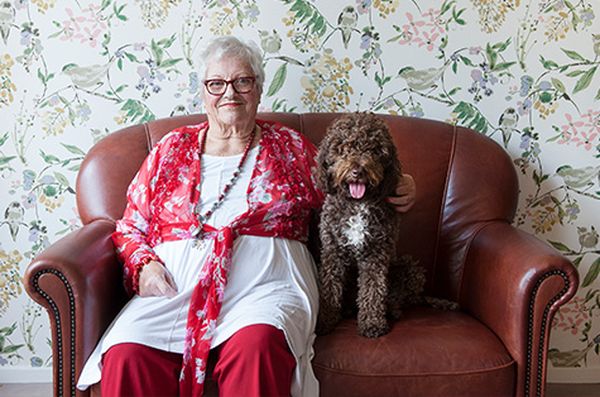 Oudere vrouw in rood-witte kleding op een leren bank met een gekrulde hond, tegen bloemenbehang. De troost van huisdieren is belangrijk in de dementiezorg.
