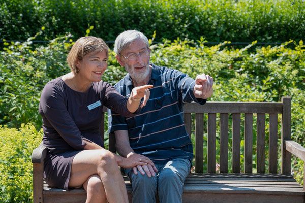 Een vrouw en een oudere man zitten op een houten bankje in een tuin; ze wijzen en glimlachen allebei naar iets in de verte. Dit moment van rust en verbondenheid benadrukt het belang van persoonlijke aandacht binnen de dementiezorg