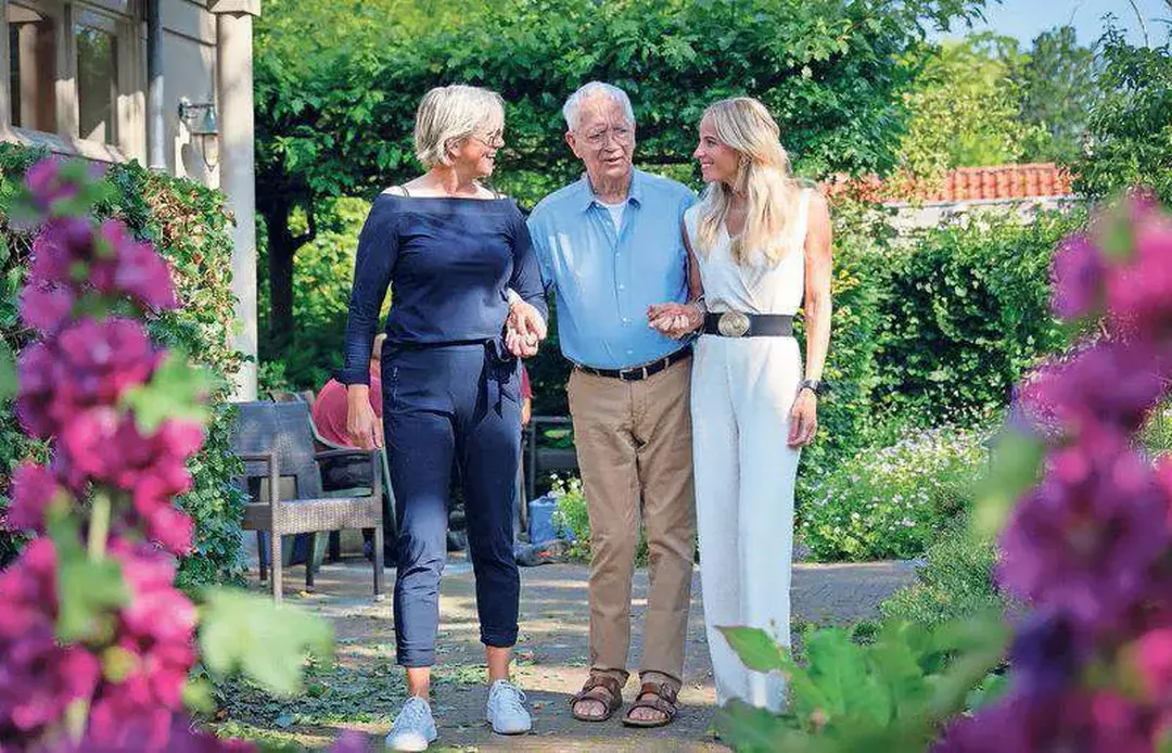 Three people walking in a lush garden, surrounded by colorful flowers and greenery, on a sunny day.