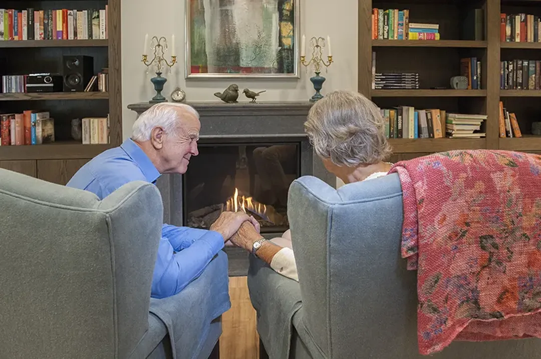 Hand in hand bij de open haard, omringd door boeken en een deken, zit een ouder echtpaar in fauteuils. Zo'n sfeer van geborgenheid is essentieel binnen de dementiezorg.