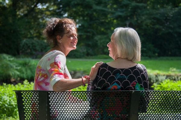 Twee vrouwen zitten lachend en pratend op een bankje in een park, omringd door groen en zonlicht. Dergelijke momenten van sociale betrokkenheid zijn essentieel binnen de dementiezorg.