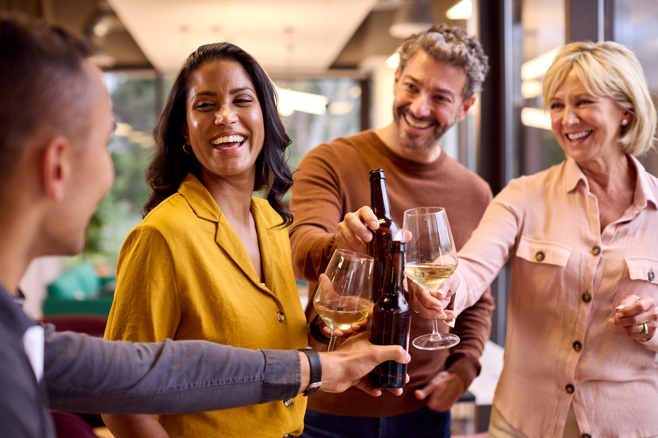 Four people cheerfully clinking wine glasses and beer bottles at a gathering, smiling and enjoying themselves indoors.