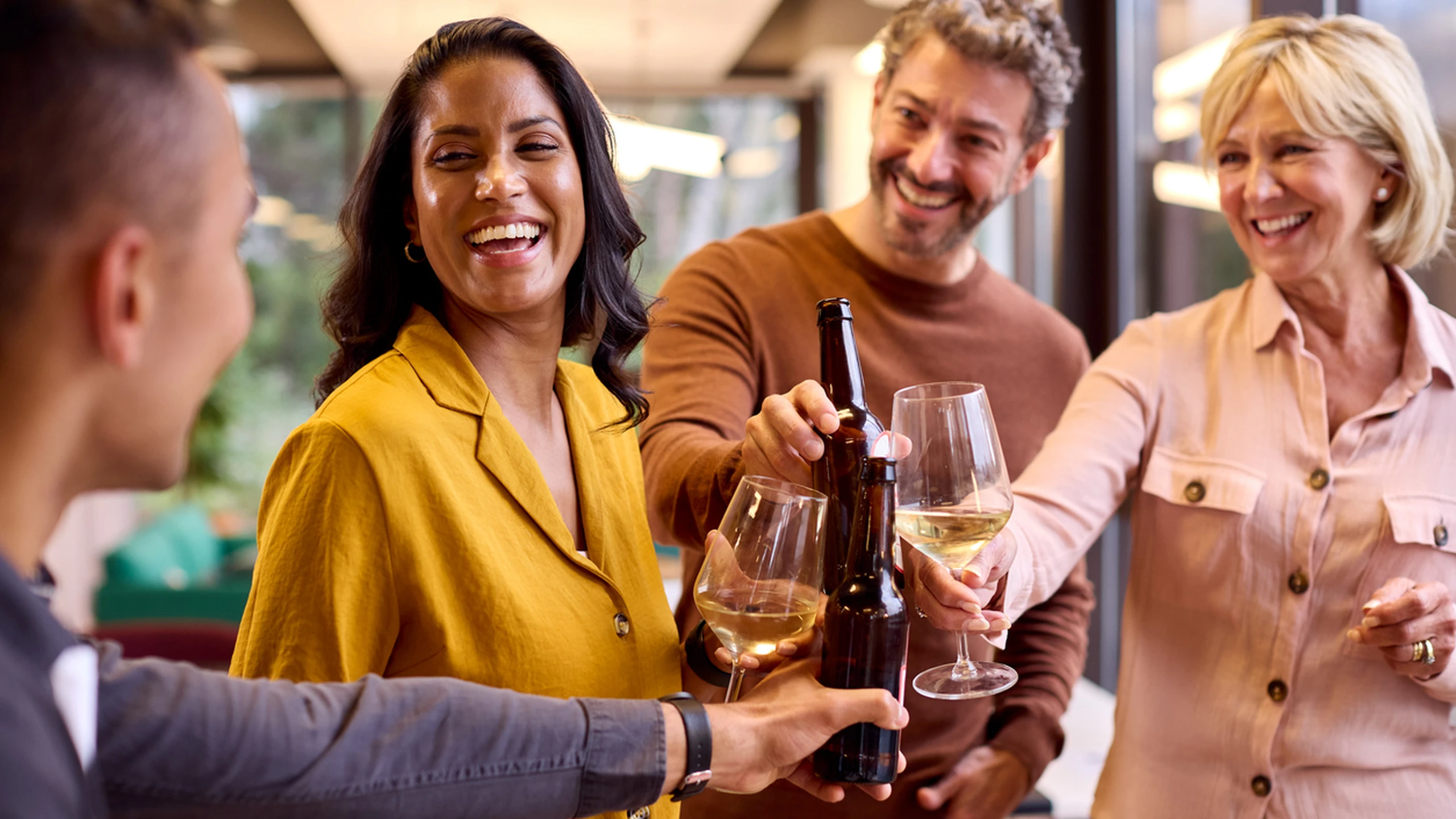 Four people cheerfully clinking wine glasses and beer bottles at a gathering, smiling and enjoying themselves indoors.