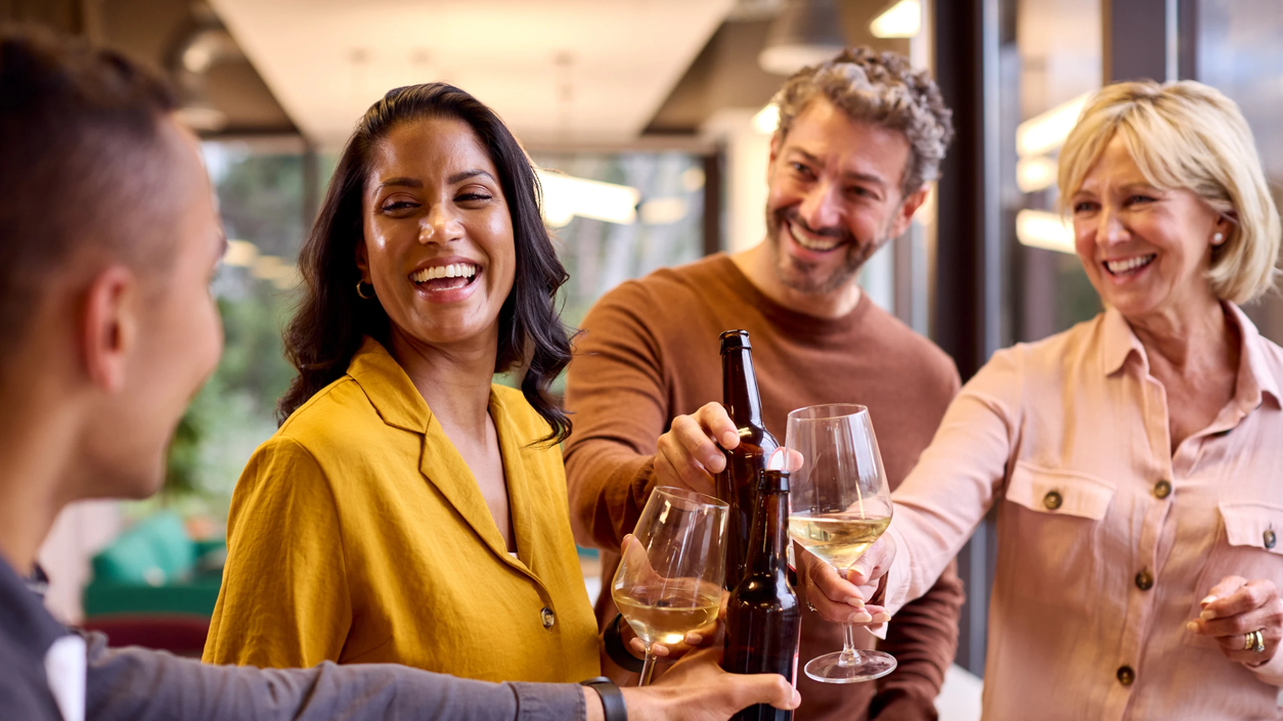 Four people cheerfully clinking wine glasses and beer bottles at a gathering, smiling and enjoying themselves indoors.