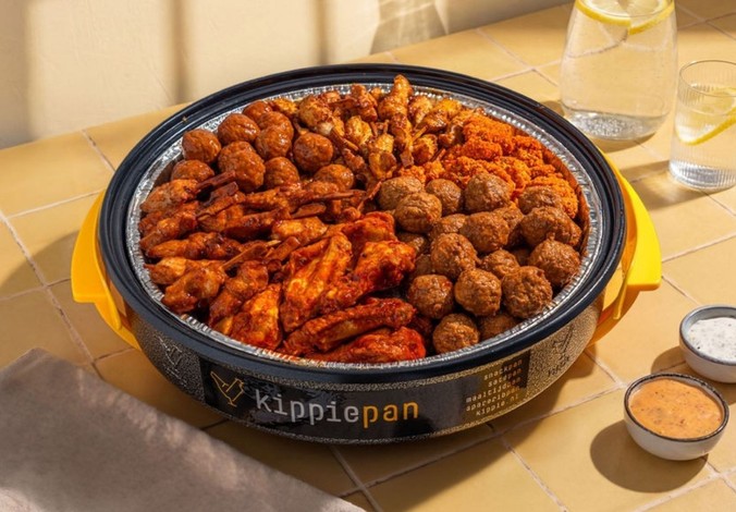 Een Kippiepan gevuld met kipkluifjes, kipgehaktballetjes, mini saté en kippeling.  Geserveerd met twee dipsausjes, karaf met water en citroen en een glas. Het is een gele ondergrond van een tafelblad