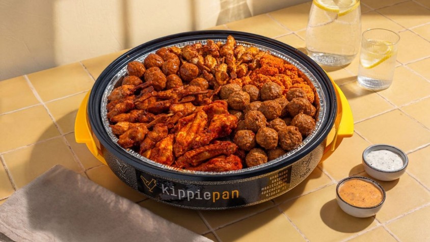 Een Kippiepan gevuld met kipkluifjes, kipgehaktballetjes, mini saté en kippeling.  Geserveerd met twee dipsausjes, karaf met water en citroen en een glas. Het is een gele ondergrond van een tafelblad