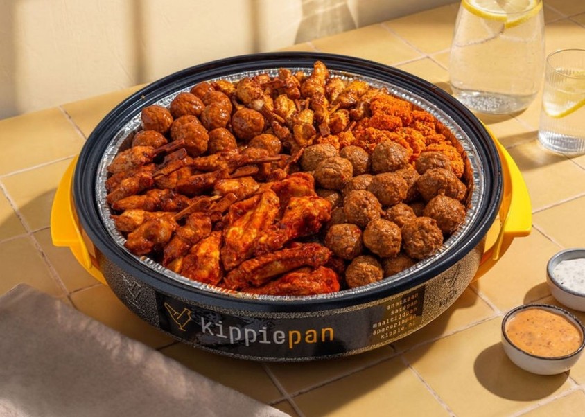 Een Kippiepan gevuld met kipkluifjes, kipgehaktballetjes, mini saté en kippeling.  Geserveerd met twee dipsausjes, karaf met water en citroen en een glas. Het is een gele ondergrond van een tafelblad